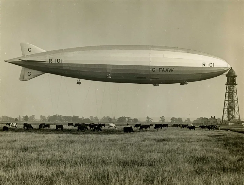 Black-and-white photo of the long R101 airship tethered to a tall mooring tower above a field of cattle